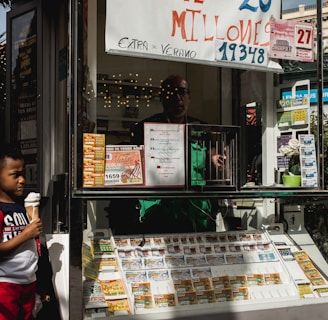 A child holding an ice cream cone stands outside a small kiosk or lottery ticket booth. The booth displays various colorful lottery tickets in the window. There are signs advertising a significant lottery prize amount in Spanish. A person can be seen partially obscured inside the booth, and plants are visible around the area, suggesting an outdoor setting.