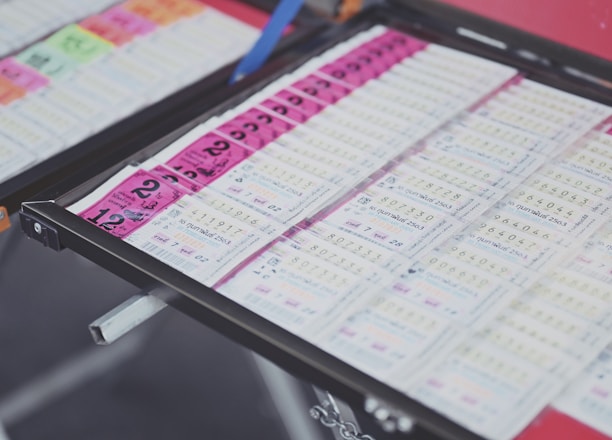 A collection of lottery tickets is organized neatly in a metal tray. The tickets display various numbers and have color-coded sections, primarily in pink and white. Some tickets have vibrant green sections, adding contrast to the overall layout.
