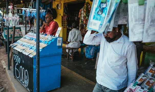 A street vendor stall displaying an array of lottery tickets clipped to a bright blue kiosk. A man in a red shirt leans on the counter, appearing thoughtful, while another man in a white shirt adjusts his hat and walks by. A variety of products hangs from strings above, and a colorful display fills the background with vibrant signs and packets. The scene suggests a busy, lively marketplace.